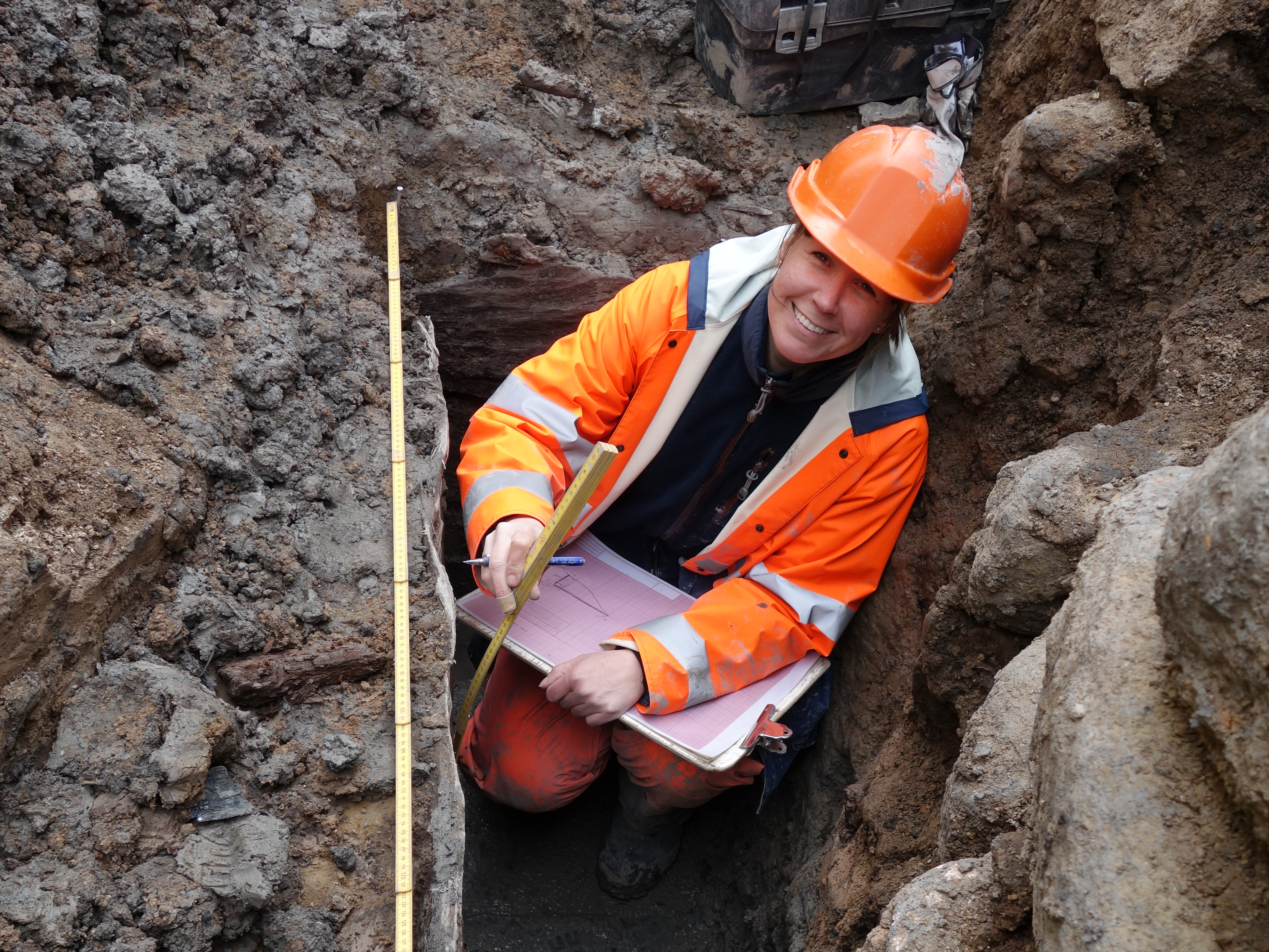 En arkeolog i full färd med att dokumentera något under en arkeologisk utgrävning i lerig, stenig grop.
