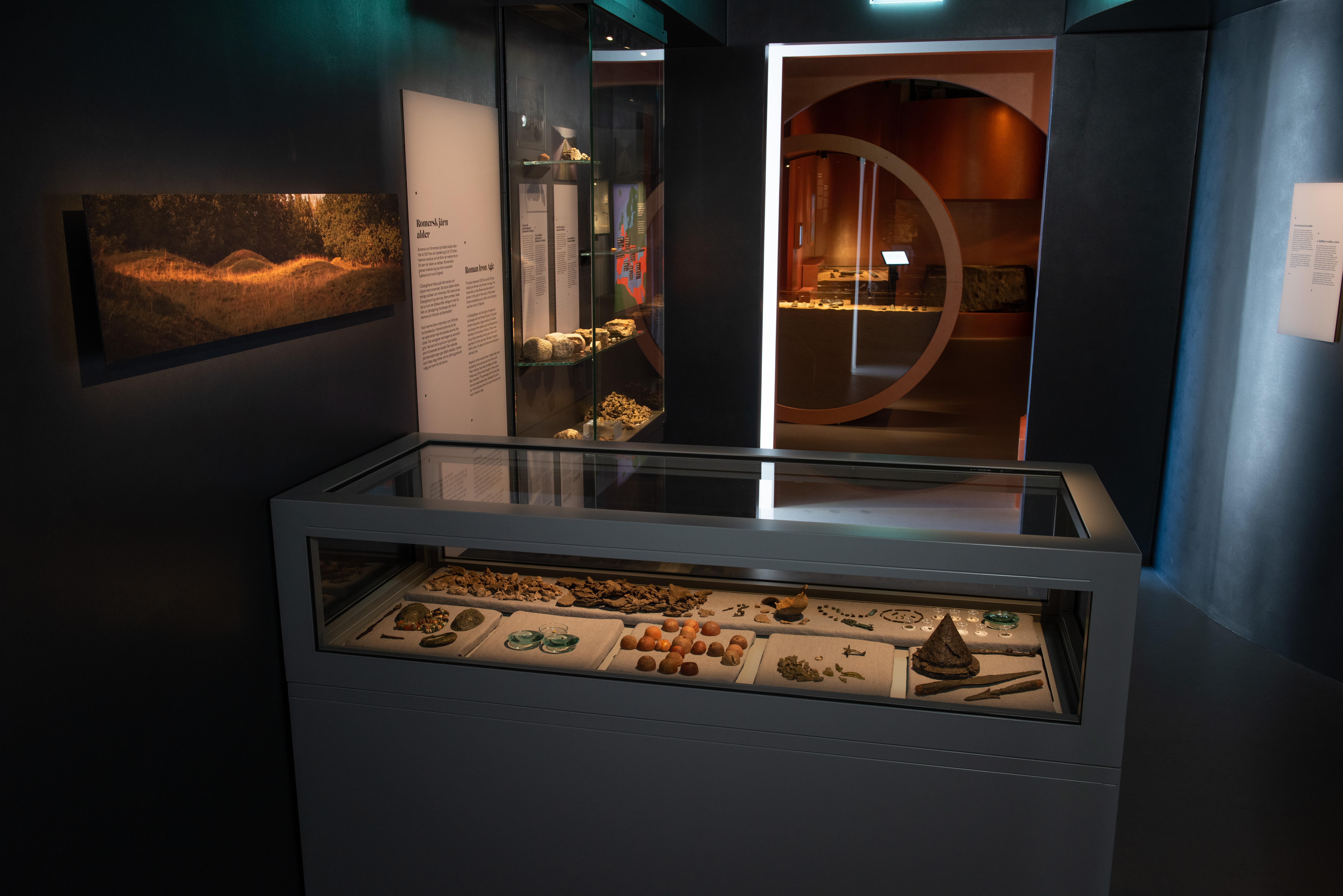 Ett rum som visar föremål från järnålder i utställningen Vägen hit. I förgrunden syns en rektangulär monter med glas på toppen. Inuti syns många arkeologiska fynd. 