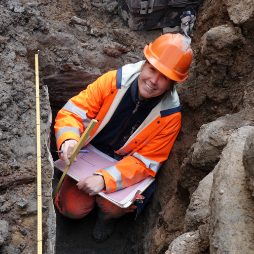 En arkeolog i full färd med att dokumentera något under en arkeologisk utgrävning i lerig, stenig grop.