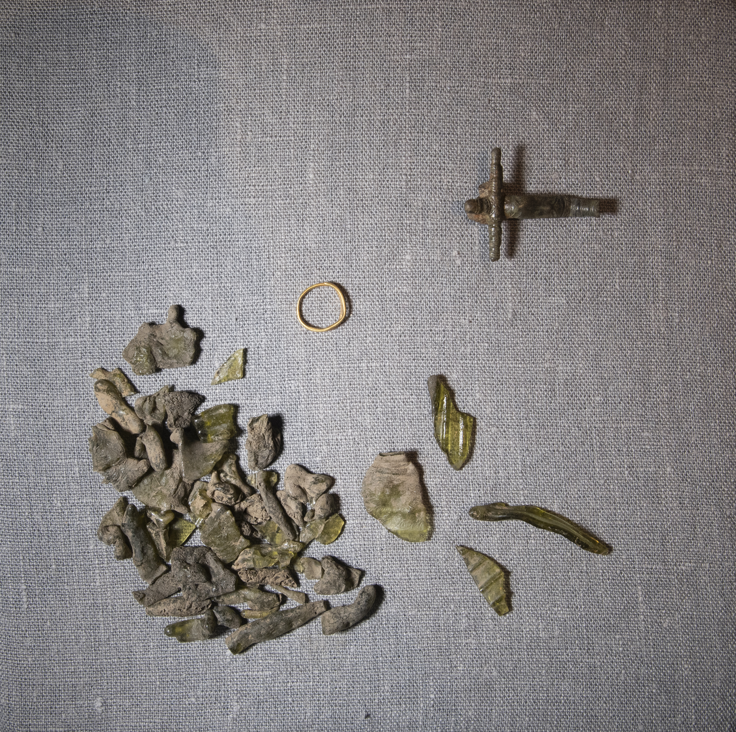 Olika fynd som hittats vid arkeologisk utgrävning, bland annat en ring, ihopsmält glasfragment och ett kors.