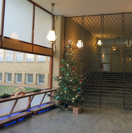 Klädd julgran i museets foajé. Under granen ligger några paket. I ett fönster syns en tänd adventsljusstake.