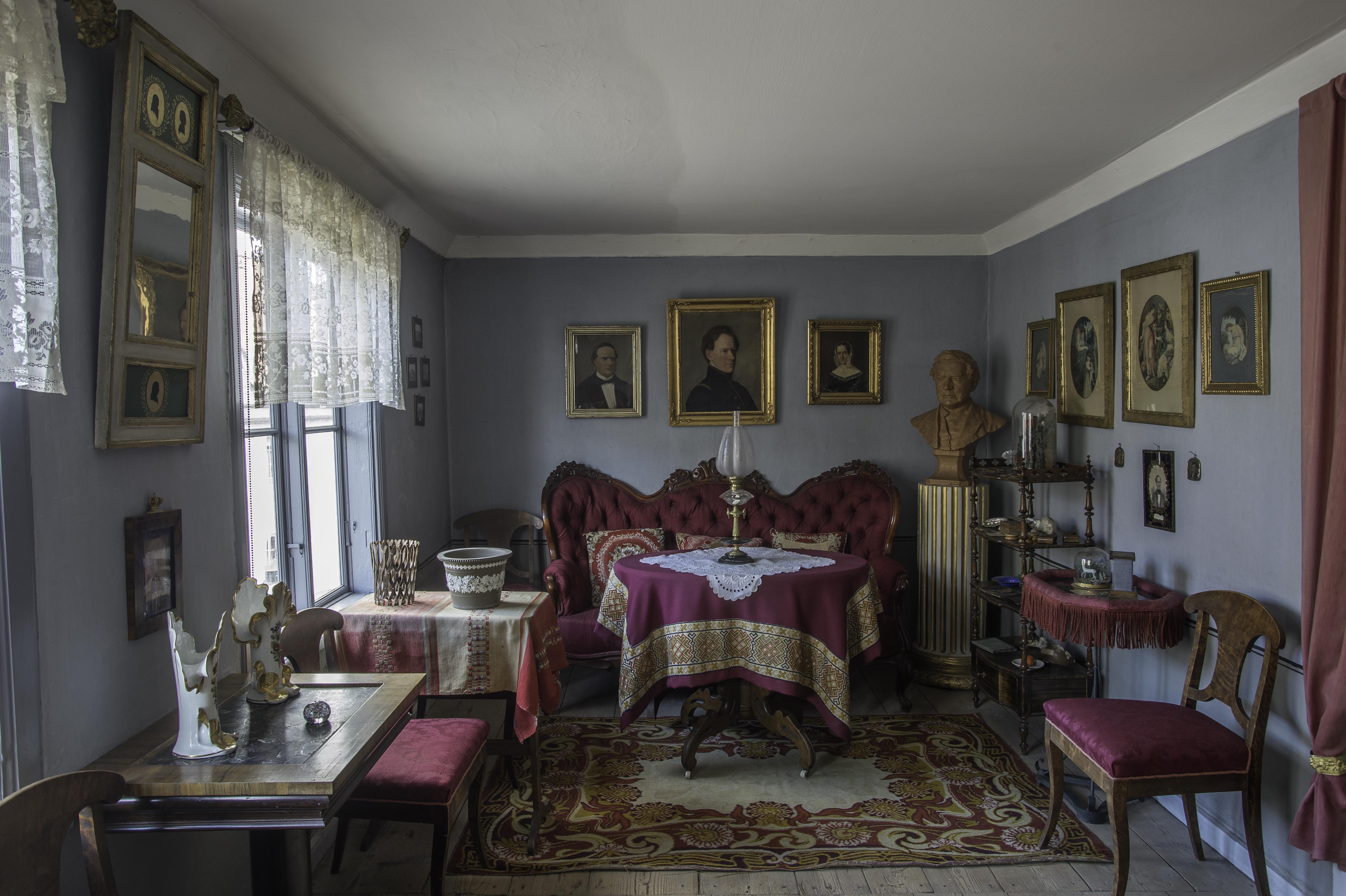 Interiör i museilägenheten i Onkel Adamsgården med möbler klädda i lila tyg, en spegel och ett flertal tavlor på väggarna, fönster med spetsgardiner och en piedestal med byst föreställande  Carl Anton Wetterbergh, pseudonymen Onkel Adam. 