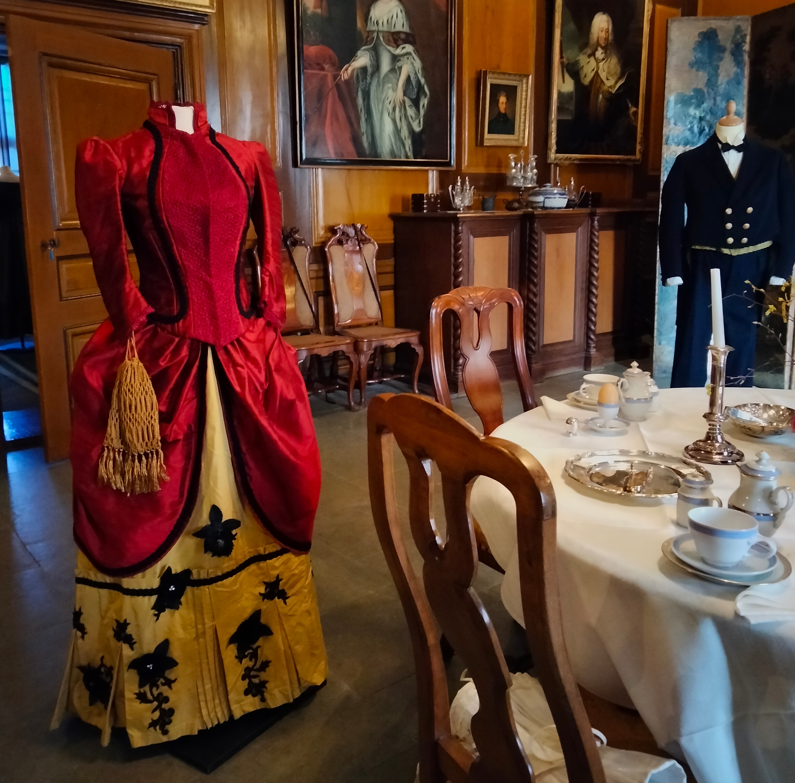 Röd klänning, ca 1880-tal, är skänkt och sydd av Tove Bjerg. I bakgrunden fröken Emilie Pipers betjänt Vilhelm Erikssons arbetskläder i original, från sekelskiftet 18-1900