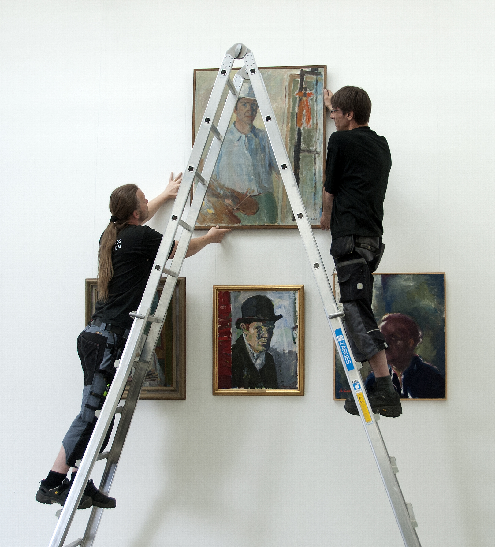 Två män på en stege hänger upp konstverk på en vägg