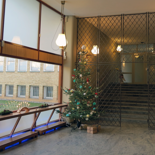 Klädd julgran i museets foajé. Under granen ligger några paket. I ett fönster syns en tänd adventsljusstake.