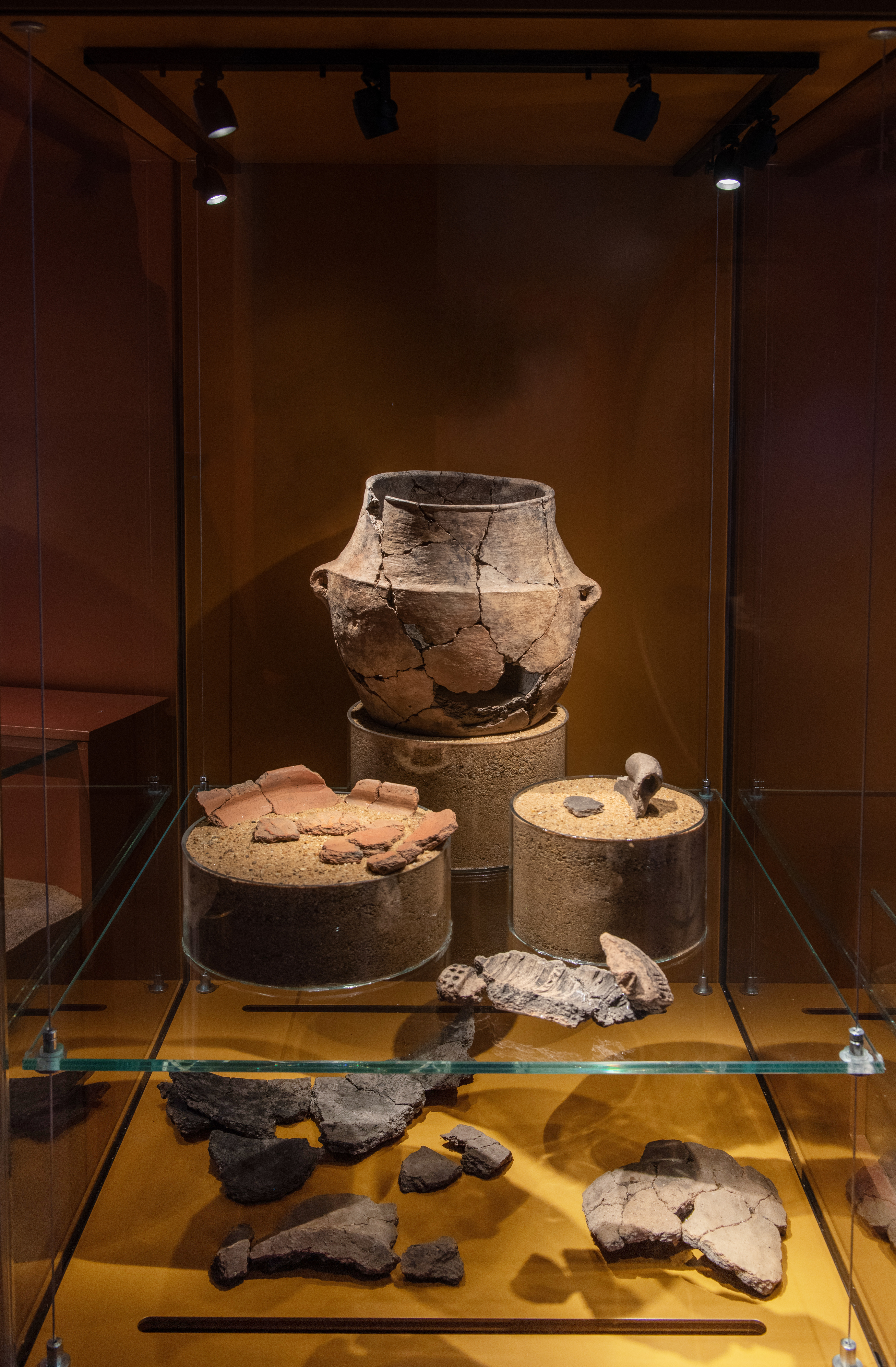 Flera arkeologiska fynd i form av keramikfragment. Uppställda på botten i en monter samt på en glashylla i samma monter. Ett av fynden visar en nästan komplett urna vars trasiga delar har limmats samman.